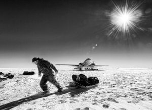 A person pulls a loaded sled across a snowy, sunlit Antarctica landscape with a propeller plane parked in the background under a radiant sun; scattered Antarctic Logistics and Expeditions gear lies on the ice.