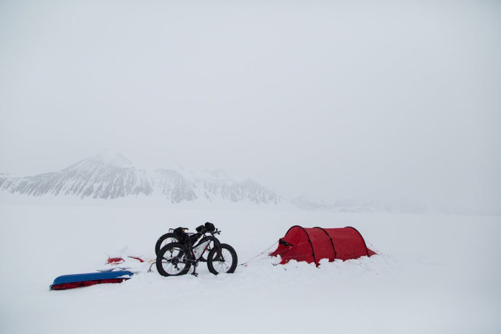 A red tent and a packed bicycle stand on a snowy landscape with sleds nearby; snow-covered mountains of Antarctica are visible in the foggy background, capturing the spirit of Antarctic Logistics and Expeditions.