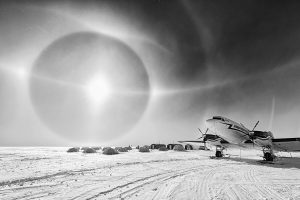 A black and white photo shows a plane on snowy ground near several tents under the bright sun, surrounded by a large halo. This remote, cold scene captures the rugged spirit of Antarctic Logistics and Expeditions in Antarctica.