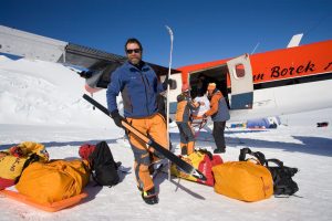 A man in orange and blue outdoor gear holds a ski and ski pole, standing by orange gear bags on snow near a small plane. Other people unload equipment from the plane in a snowy Antarctic Logistics and Expeditions setting.