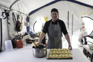 A man wearing an apron stands inside a tent kitchen in Antarctica, smiling behind a table with a large pot and a tray of baked food. Pots, utensils, and cooking supplies are visible—part of Antarctic Logistics and Expeditions at the South Pole.