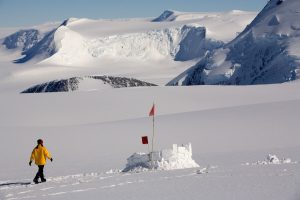 A person in a yellow jacket walks across a snowy landscape toward a small snow structure with red flags at the South Pole, with snow-covered mountains and glaciers in the background under a clear Antarctic sky.