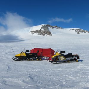 Two yellow snowmobiles are parked on a snowy field next to a red tent, with a snow-covered mountain and blue sky in the background—an iconic scene from Antarctic Logistics and Expeditions near the South Pole in Antarctica.