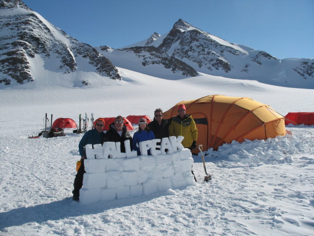Five people stand behind a snow wall labeled HALL PEAK in Antarctica’s snowy mountain landscape, with orange tents and gear from Antarctic Logistics and Expeditions in the background under a clear blue sky.