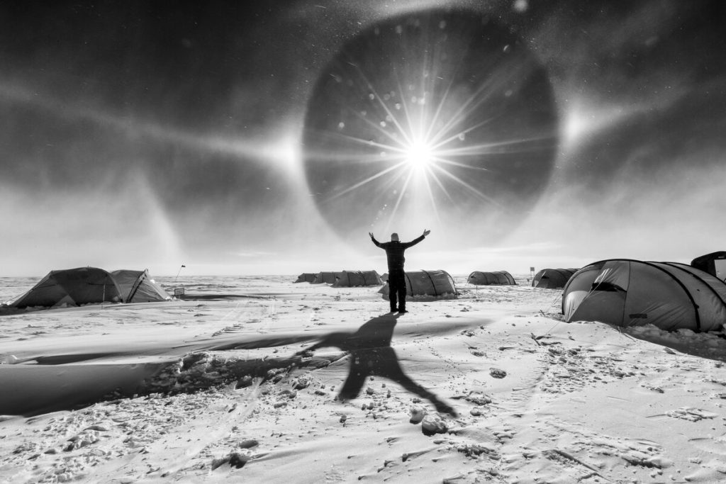 A person stands with arms raised on a snowy landscape at the South Pole, surrounded by tents. The sun shines brightly overhead, creating a dramatic halo effect and casting a long shadow on the snow. Black and white photo.