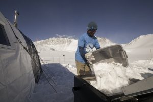 A person wearing sunglasses and winter clothing empties a container of snow into a large bin beside a tent in Antarctica’s snowy, mountainous landscape under a clear blue sky.