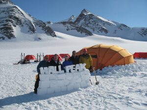 Five people stand behind a snow wall labeled HALL PEAK in Antarctica’s snowy mountain landscape, with orange tents and gear from Antarctic Logistics and Expeditions in the background under a clear blue sky.