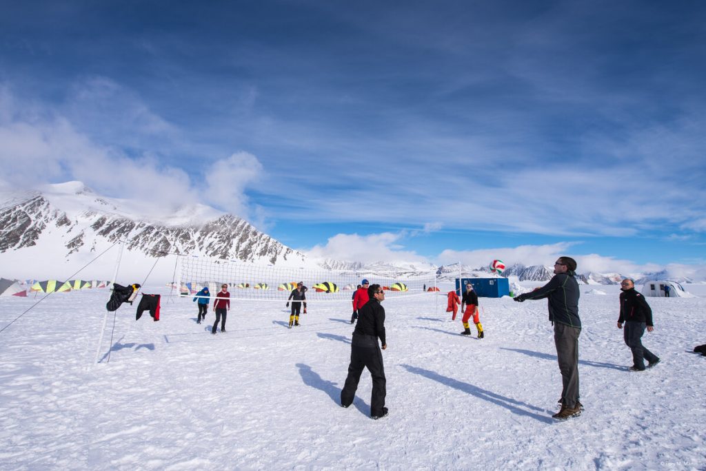 A group of people dressed in winter gear play volleyball on a snowy field in Antarctica, with tents and snow-covered mountains in the background under a blue sky.