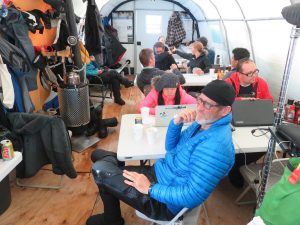 People in winter gear work and socialize inside an Antarctic tent, seated at tables with laptops, drinks, and papers. Coats and gear hang on the walls. The atmosphere appears busy and collaborative at the South Pole.