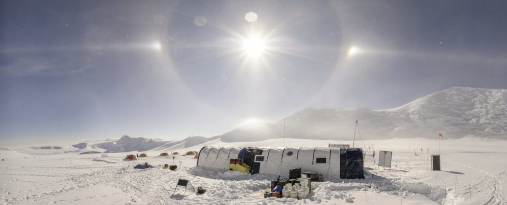 A snowy Antarctic research camp with tents and equipment under a bright sun halo, surrounded by snow-covered mountains and a clear sky—an iconic scene in Antarctica supported by Antarctic Logistics and Expeditions.