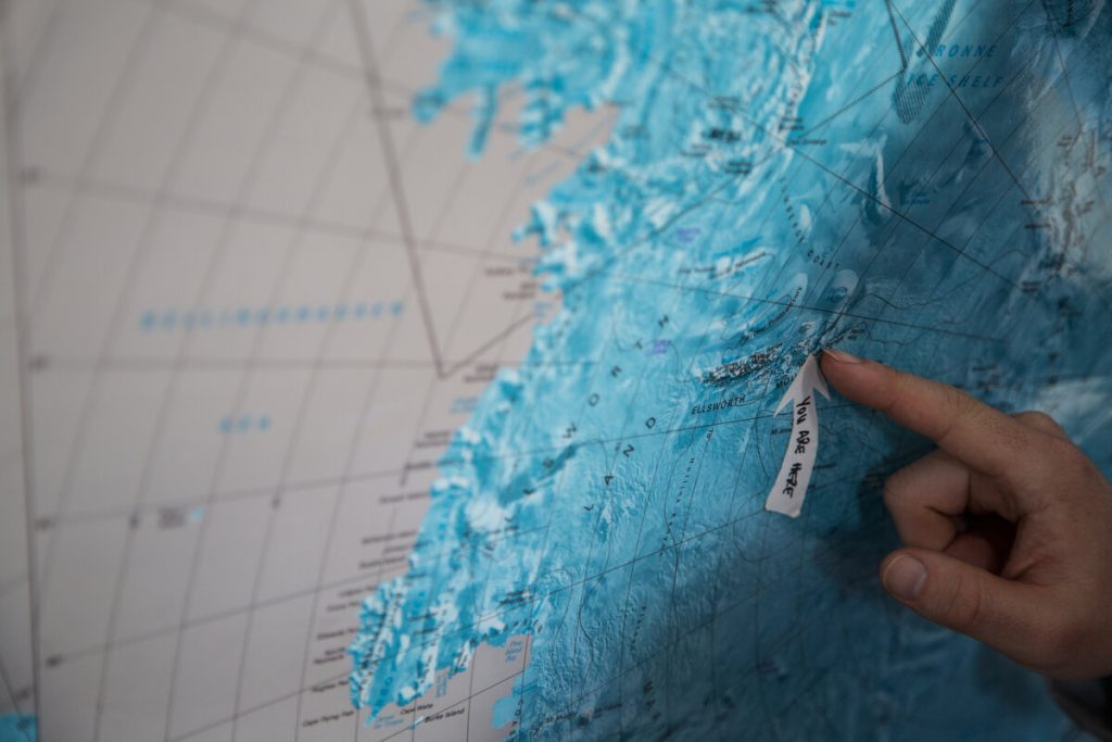 A hand points to a location labeled You Are Here on a blue-toned topographic map of Antarctica, indicating a specific spot among various geographic features and lines near the South Pole.