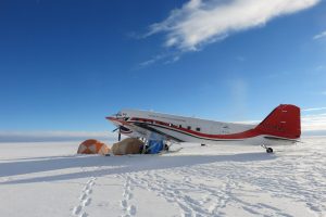 A red and white airplane is parked on snowy ground next to two orange tents under a clear blue sky, with snow and ice stretching to the horizon—an iconic scene from Antarctic Logistics and Expeditions in Antarctica.