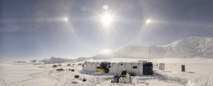 A snowy research camp with tents stands under a bright sun halo in the vast icy landscape of Antarctica. Mountains and additional tents, set up by Antarctic Logistics and Expeditions, are visible in the background under a clear sky.