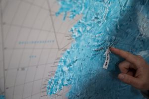 A hand points to a label reading YOU ARE HERE on a blue-toned map, highlighting a specific location along an icy coastline near Antarctica, possibly marking an Antarctic Logistics and Expeditions route.