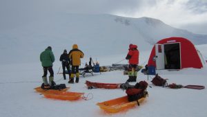 Four people in winter gear stand on snow near orange sleds and equipment outside a red tent, with snowy mountains in the background under a cloudy sky, capturing the spirit of Antarctic Logistics and Expeditions in Antarctica.