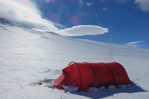 A bright red tunnel tent stands on a snowy, windswept Antarctic landscape under a blue sky with wispy clouds; ski poles and gear are placed beside the tent—a scene reminiscent of South Pole expeditions.
