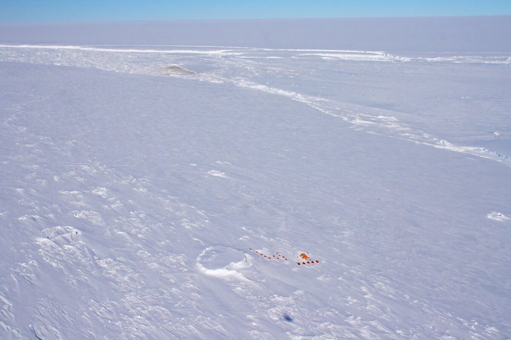A vast, snow-covered landscape in Antarctica with a small cluster of orange tents near the bottom center, surrounded by icy ridges under a clear blue sky.