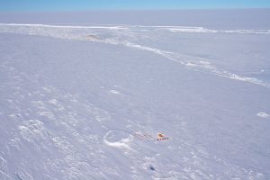 A vast, snow-covered landscape in Antarctica with a small cluster of orange tents near the bottom center, surrounded by icy ridges under a clear blue sky.