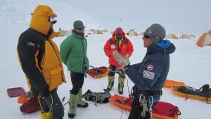 Four people in winter gear stand on snow near orange tents, deep in discussion. Sleds and equipment lie nearby, with one gesturing while holding an object—an Antarctic Logistics and Expeditions team operating in a cold, mountainous environment.