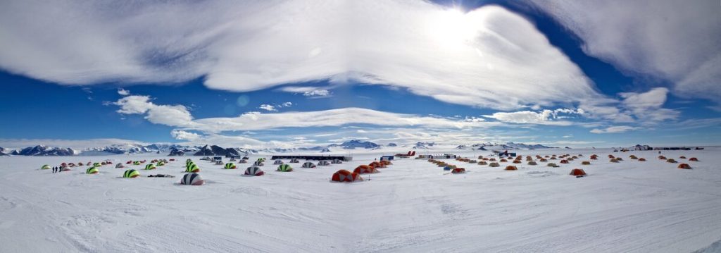 A wide snowy landscape under a bright sky, scattered with colorful tents arranged across the ice in Antarctica. Mountains are visible in the distance as the sun shines through wispy clouds, capturing the spirit of Antarctic Logistics and Expeditions.