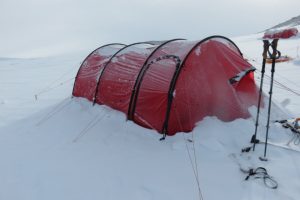 A red tunnel tent is partially buried in snow on a snowy landscape near the South Pole. Trekking poles and equipment are visible nearby, highlighting a cold and remote camping location supported by Antarctic Logistics and Expeditions.