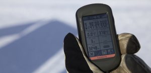 A gloved hand holds a Garmin GPS device displaying coordinates, altitude, speed, and battery information against a snowy Antarctic Logistics and Expeditions backdrop.