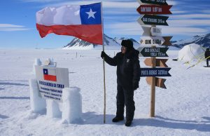 A person in winter clothing holds a Chilean flag on a snowy landscape near a signpost with distances to various locations at the South Pole in Antarctica. Mountains and a clear sky are in the background.