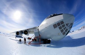 A large cargo plane parked on a snowy, icy landscape in Antarctica under a bright sun, with mountains in the background. People stand near the plane, and snowmobiles—part of Antarctic Logistics and Expeditions—are visible on the ice.
