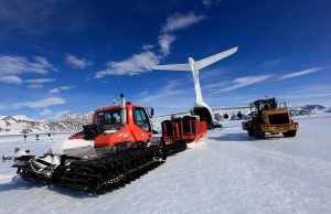 A red snowcat and a yellow loader, part of Antarctic Logistics and Expeditions, unload cargo from a large airplane on an icy runway in Antarctica’s mountainous, polar region under a blue sky with scattered clouds.