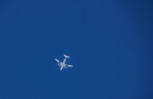 A white airplane operated by Antarctic Logistics and Expeditions is flying high in a clear blue sky over Antarctica, viewed from below with no clouds visible.