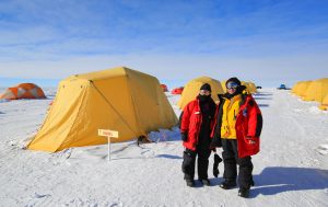 Two people in heavy winter gear stand on snow beside a yellow tent at an Antarctic Logistics and Expeditions camp filled with similar tents under a blue sky. A small sign reading Häggel is visible in front of the tent.