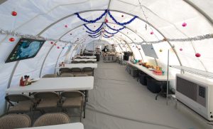 A large white tent at the South Pole serves as a dining area with long tables and chairs, decorated with tinsel, garlands, and ornaments. Food and supplies line side tables, and maps of Antarctica are mounted on the walls.