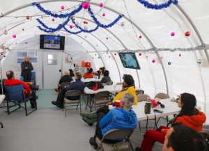 A group of people sit on chairs and at tables inside a decorated white tent at the South Pole, listening to a speaker at the front. Tinsel and ornaments hang from the ceiling, while Antarctic Logistics and Expeditions images display on screens.