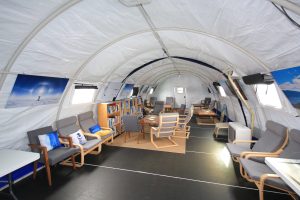 A spacious, well-lit tent with arched white walls, furnished with armchairs, tables, bookshelves, and framed photos. This cozy lounge offers a relaxing reading area for those on an Antarctic Logistics and Expeditions journey to the South Pole.