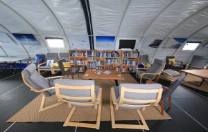 A cozy reading area with gray-cushioned chairs arranged in a circle around a wooden table, surrounded by bookshelves, inside a dome-shaped room reminiscent of Antarctic Logistics and Expeditions spaces near the South Pole.