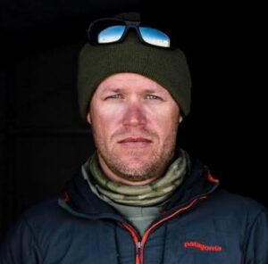 A man wearing a green beanie with sunglasses on top, a patterned neck gaiter, and a dark jacket stands against a dark background, evoking the spirit of Antarctic Logistics and Expeditions at the South Pole.