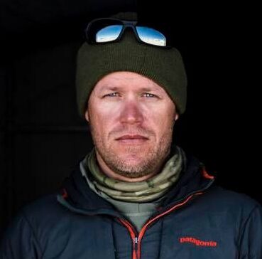 A man wearing a green beanie with sunglasses on top, a patterned neck gaiter, and a dark jacket stands against a dark background, evoking the spirit of Antarctic Logistics and Expeditions at the South Pole.