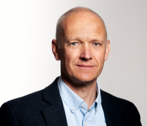A middle-aged man with short, light hair is wearing a dark blazer over a light blue button-up shirt, standing against a plain, light background—ready for his next Antarctic Logistics and Expeditions journey.