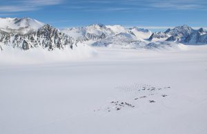 A remote research camp operated by Antarctic Logistics and Expeditions with tents, vehicles, and small structures sits on a vast snowy plain in Antarctica, surrounded by snow-covered mountains under a clear blue sky.