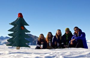 Five people sit in the snow, smiling, next to a wooden Christmas tree with a red hat. Snowy mountains and a clear blue Antarctic sky frame the adventure with Antarctic Logistics and Expeditions in the background.