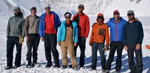 Eight people dressed in winter outdoor clothing stand side by side on snow with a snowy mountain backdrop. Some wear sunglasses and hats, and all are smiling or posing for the photo.