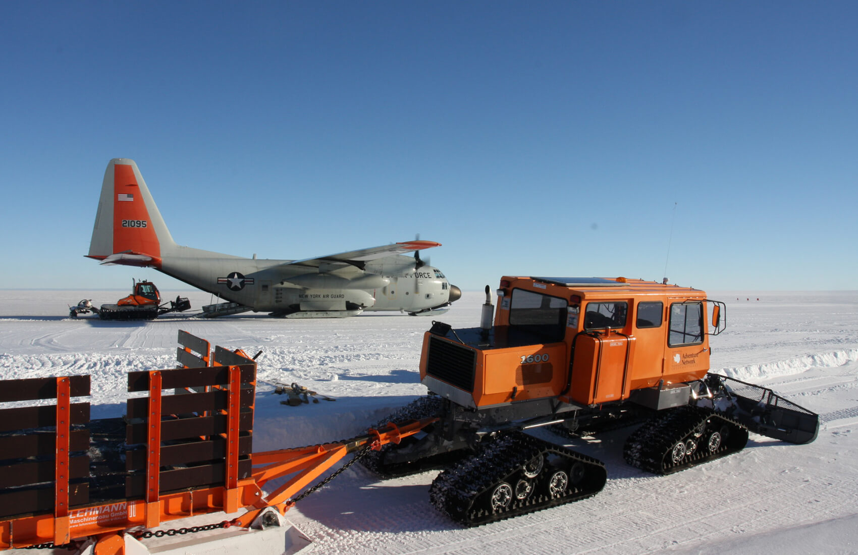 An orange snowcat vehicle towing cargo on snow, with another snowcat and a U.S. military transport plane in the background at an Antarctic Logistics and Expeditions site on the flat, snowy landscape of Antarctica under a clear blue sky.