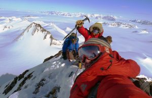 Three climbers in red and blue winter gear stand on a snowy Antarctic ridge, surrounded by pristine peaks. One holds an ice axe aloft while another takes a selfie with reflective goggles—a classic moment from an Antarctic Logistics and Expeditions adventure.