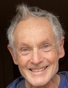 An older man with light skin, short gray hair, and blue eyes smiles warmly at the camera. Wearing a blue shirt, he stands against a dark background—his gaze hinting at past adventures in Antarctica with Antarctic Logistics and Expeditions.