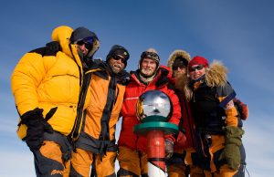 Five people in heavy winter gear stand close together at the South Pole, smiling around a reflective silver sphere atop a red and white pole, with a blue Antarctic sky in the background.