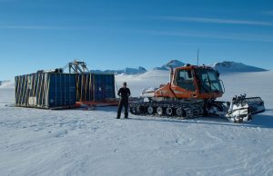 A person stands next to an orange tracked snow vehicle pulling a large, container-like structure across a snowy, mountainous Antarctic landscape under a clear blue sky—showcasing the challenges of Antarctic Logistics and Expeditions.