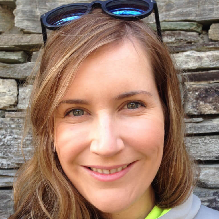 A woman with long light brown hair smiles at the camera, wearing sunglasses on her head. She stands in front of a stone wall in natural light, looking ready for new adventures—perhaps even with Antarctic Logistics and Expeditions.
