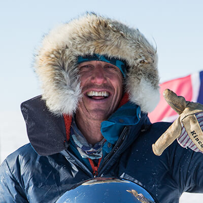A smiling person wearing a fur-lined hooded parka and gloves stands outdoors in snowy Antarctic conditions, with a flag in the background and bright sunlight shining—capturing the spirit of Antarctic Logistics and Expeditions.