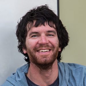A man with wavy dark hair and a beard smiles at the camera. Wearing a blue hoodie over a dark shirt, he stands against a light background, ready for his next adventure with Antarctic Logistics and Expeditions in Antarctica.