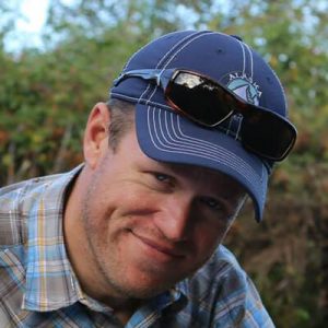 A man in a blue plaid shirt and blue baseball cap with sunglasses resting on top smiles at the camera outdoors, surrounded by greenery—a far cry from the icy landscapes managed by Antarctic Logistics and Expeditions.